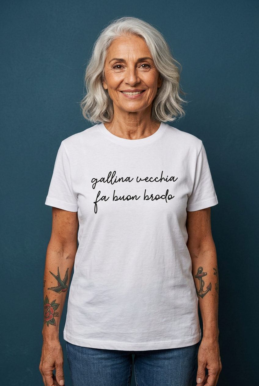 Modella over 60 con capelli argentati e tatuaggi old school indossa la T-shirt Gallina vecchia fa buon brodo su sfondo blu petrolio